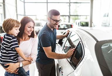 A family of 3 smiling in a dealership looking at new vehicles.