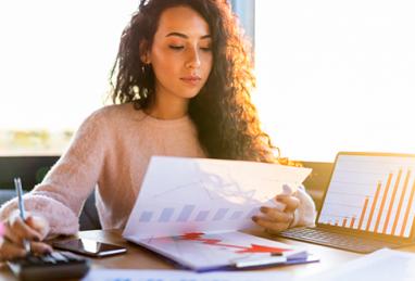 young woman sitting at table reviewing her investment portfolio, laptop, documents, investment reports, calculator, investing, wealth, building wealth