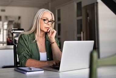 older woman with long gray hair sitting at table using laptop, online activity, online security, digital banking, fraud, scam, cybersecurity, account protection, tips