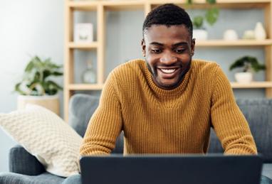 young black man sitting on couch at home smiling and using laptop, reviewing finances, creating a budget, budgeting, budgeting for the new year