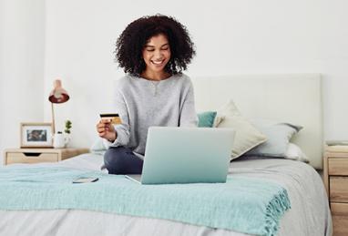 young African-American woman sitting on her bed at home holding credit card and using laptop, credit, building credit, credit card, credit report, credit score, online shopping