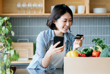 young Asian woman holding credit card and smartphone while standing in her kitchen, credit card, groceries, fresh produce, kitchen, at home