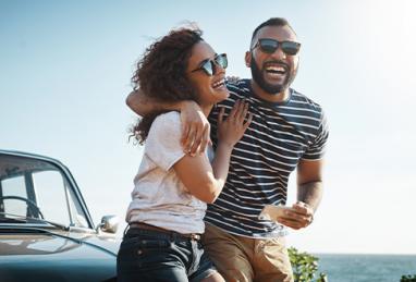 happy young couple smiling and laughing beside car in front of sky and water on the horizon, summer skies, road trip, car