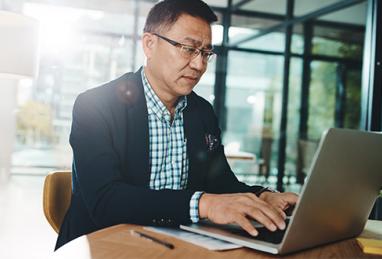 older adult Asian man sitting at table typing on laptop