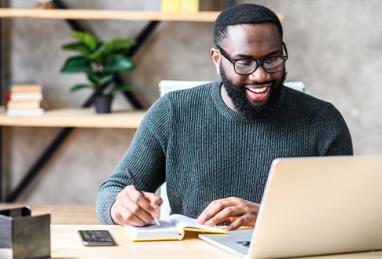 black man sitting at desk using laptop and writing in notebook, using a calculator, creating a budget, budgeting